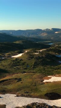 Vertical Screen: Beartooth Highway in Montana USA reveals stunning alpine meadows with scattered snow patches and pristine mountain lakes. Drone captures golden hour light across vast wilderness.