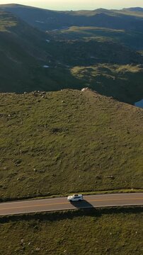 Vertical Screen: Beartooth Highway in Montana USA features white SUV traversing scenic mountain road. Aerial drone view captures alpine tundra landscape with distant peaks and lake at golden hour.