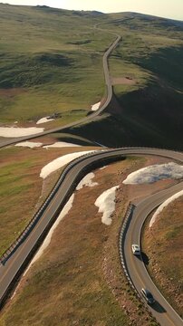Vertical Screen: Beartooth Highway in Montana USA winds through alpine terrain with snow patches and green meadows as white car navigates hairpin curves during golden hour light.