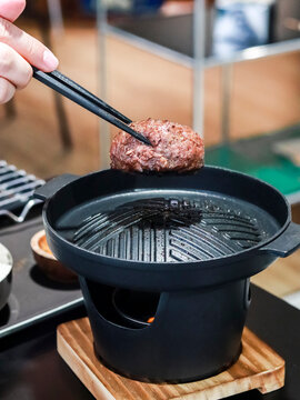 A close-up shot of a hand using chopsticks to hold a thick, juicy Japanese wagyu hamburger steak (Hambagu) over a hot, smoking personal tabletop cast iron grill. 