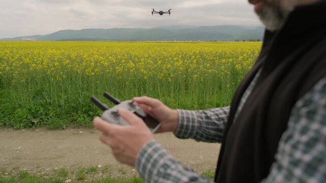 Moody and worried agronomist pilot operating drone for inspection over vast yellow rapeseed field under overcast sky, dynamic pull away tracking shot, 4k