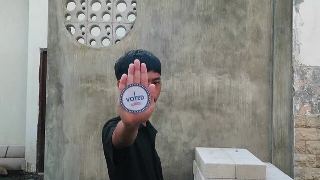 A young man holds up a 'us vote' sticker in front of his face standing in a room with a concrete wall and decorative circular pattern