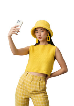 Young east asian woman in lemon-yellow ensemble and bucket hat makes a duck face with a smartphone, posing for a selfie in a minimalist studio, embodying trendy gen-z self-expression