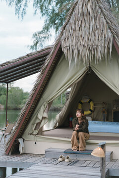 Glamping nature retreat young Asian woman relaxing sitting on porch of thatched roof tent over lake water concept of slow living solo travel weekend getaway mindfulness digital detox and eco-tourism
