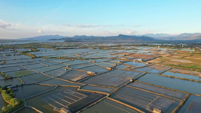 Aerial drone footage of a vast salt pond field valley, with villages, huts, in Woha area, Sumbawa island, Indonesia, at golden sunset, with Bima airport behind and mountains around the city