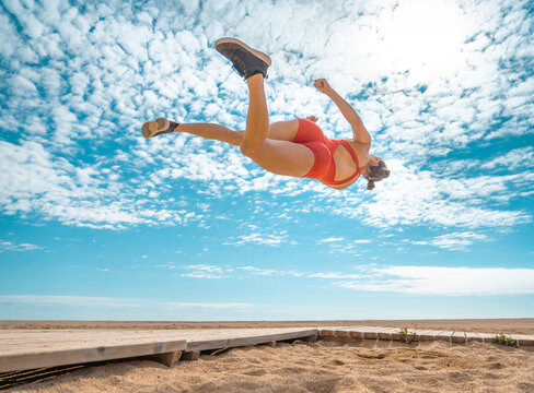 Athletic young woman, in a tight red jumpsuit, performing a high webster 360 flip mid-air above a beach boardwalk.