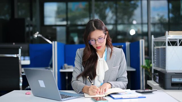 A young professional Asian businesswoman smiling while using her smartphone and working with a laptop at her office