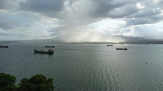 Aerial close up drone footage of Bima city harbor bay area at sunset with heavy clouds and rain, in Sumbawa island, Indonesia, with many tanker ships anchored, and Doro Oromboha mountain in background