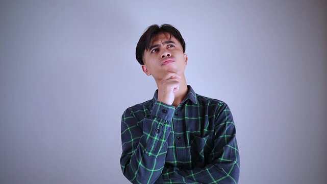 Thoughtful young asian man looking up holding chin with concentrated expression thinking hard about business solution against white studio background
