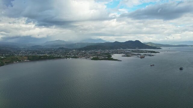 Aerial drone footage of Bima city harbor bay area at sunset with heavy clouds and rain, in Sumbawa island, Indonesia, with many tanker ships anchored, and the whole city and hills in the background