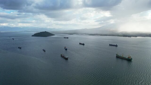 Aerial drone footage of Bima city harbor bay area at sunset with heavy clouds and rain, in Sumbawa island, Indonesia, with many tanker ships anchored, and Doro Oromboha mountain in background