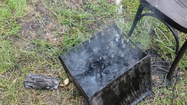 Person extinguishing smoking barbecue grill with water, pouring onto smoldering coals, first person view
