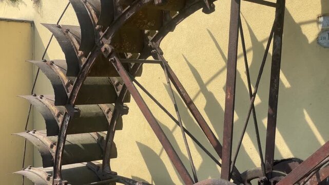Rustic Water Wheel: A weathered, industrial water wheel made of metal and wood stands against a yellow wall, casting long shadows under the bright sun. Robbiano, Italy