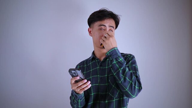 Sad and confused young Asian man holding a credit card while looking at his smartphone, experiencing financial problems, online shopping failure, or cyber scam concept.