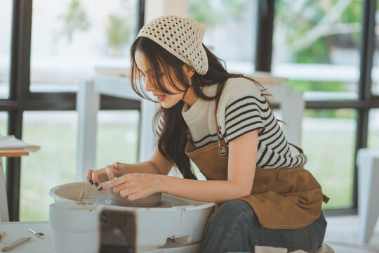 Happy asian female ceramic artist wearing apron cutting clay bowl from pottery wheel using string &ndash; traditional handmade pottery process in creative studio with focus on craft and artistic lifestyle