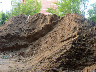 Fototapeta premium A large pile of excavated brown soil sits at a construction site. Heavy machinery tracks mark the ground at its base. In the background, green trees and a red brick building are visible under daylight