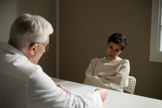 Middle aged woman sitting restrained during psychiatric evaluation, listening to senior male doctor, conveying mental health crisis and treatment. For therapy, diagnosis, care campaigns