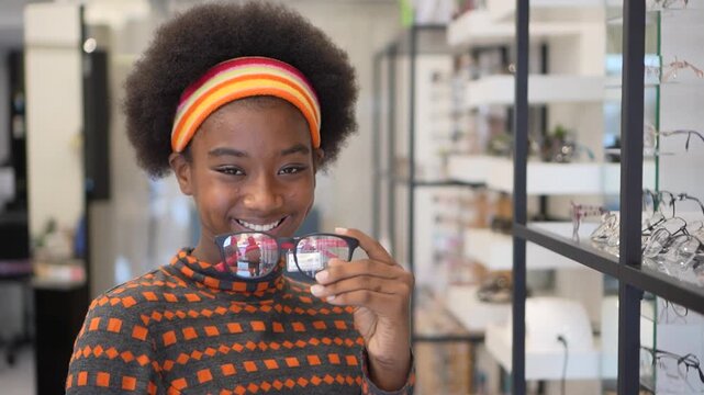 Smiling young African woman with curly afro hairstyle wearing colorful headband and glasses optical store, holding eyewear, surrounded by display shelves, showcasing modern, cheerful, confident
