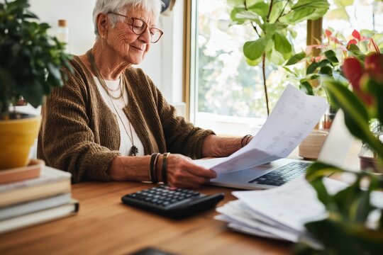 Elderly woman reviewing bills and invoices in a sunlit home office filled with plants and comfort