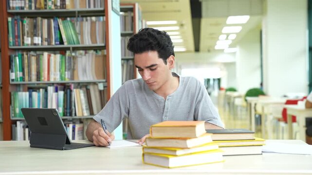 Young Caucasian male student focusing and writing on notebook with digital tablet in university library. Handsome teenage guy studying, doing homework near bookshelves in school. Education.