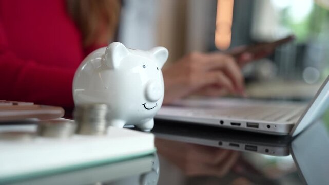 Woman using a smartphone beside a piggy bank and calculator, representing digital budgeting, saving money, and personal financial management.