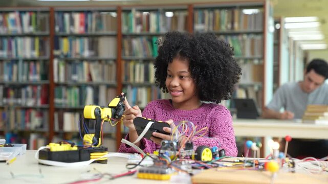 Focused young afro girl testing and controlling a robotic arm with a remote controller in library. African American student learning STEM education, robotics and engineering.