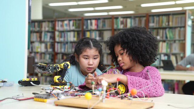 Multiethnic little schoolgirls working together on a STEM robotics project. African American and Asian children connecting wires and building an electronic robot in library. Teamwork, diversity.