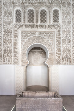 Visitors explore the prayer room at Madrasa Ben Youssef. The room features intricate designs on the walls and a central fountain. It showcases traditional architecture and cultural significance.
