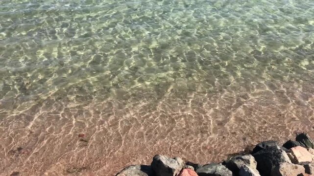 Clear shallow sea lapping rocky shore, transparent aquamarine water revealing pebbled seabed and granite boulders, sunlit ripples