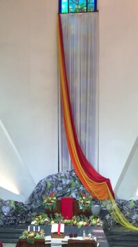 A vibrant altar draped with flowing red and yellow fabric, set against a backdrop of a stained-glass window and rocky landscape.