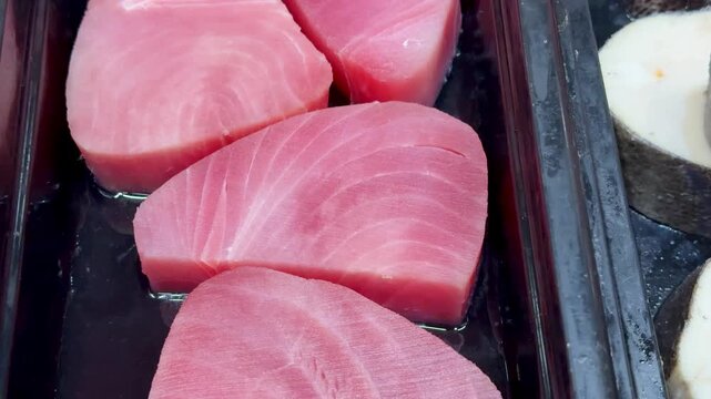 Pink tuna steaks on black tray, supermarket shelf closeup, chilled display of raw fresh fillets arranged in neat rows, subtle