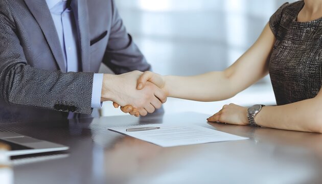 Two business professionals, a man and a woman, shaking hands over a contract on a table, symbolizing agreement and partnership.