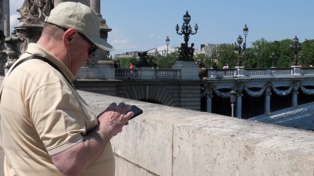 A medium shot captures a male tourist wearing a cap and sunglasses, standing
by the stone balustrade of the iconic Pont Alexandre III in Paris. He is focused
on typing and using his smartphone. 