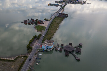 Naklejka premium Aerial image showing a narrow paved causeway flanked by unique wooden stilt structures, restaurants, and small docked boats on the mirrored waters of Patok Lagoon, Albania.