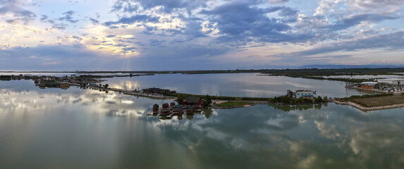 Fototapeta premium Aerial panorama capturing clusters of wooden stilt cabins along a causeway, with a dynamic sunset sky mirroring onto the tranquil waters of Patok Lagoon, Albania.