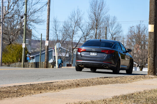 Quebec, Canada, 26 April 2026 : Dark gray Kia Optima sedan drives away on a street with bare trees and utility poles
