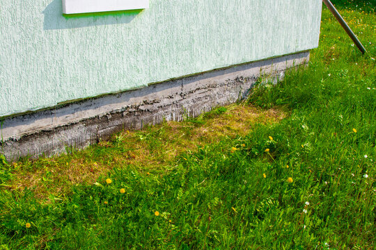 Damaged concrete house foundation with cracks and moisture weathering. Maintenance problem on residential building exterior wall and green garden lawn. Structural defect and dampness