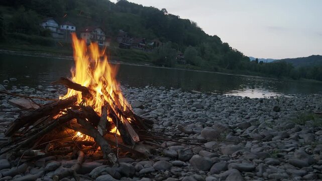 4K full-view video of a campfire placed at the far left side of the frame on a rocky bank of the Drina River at sunset, captured from Serbia with the mountains of Bosnia and Herzegovina in view.