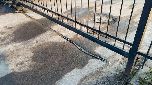 Automatic iron gate opening over asphalt parking lot, closeup of hinge and post, visible manhole cover and oil stains on pavement