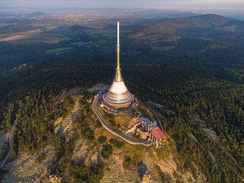 Aerial view of the iconic Jested Tower on the mountain ridge near Liberec, Czech Republic, surrounded by forests and hills during warm sunset light in a scenic natural landscape.