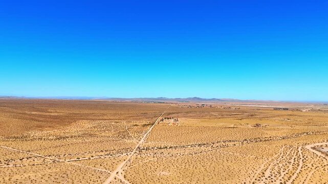 Flight over the vast desert covered by the multiple little shrubs and some ground roads. Heading to the rocks against blue clear sky in the backdrop. California, USA.