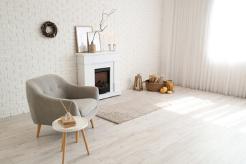 Fototapeta premium Interior of stylish room with modern fireplace, armchair and autumn decorations near white brick wall