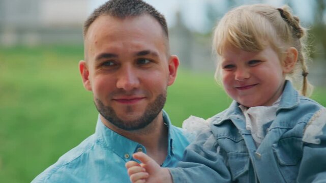 Father holds child with gentle smile. Child beams, arms raised in joyful gesture. Mother's back is to camera, capturing family moment. Soft green backdrop frames tender