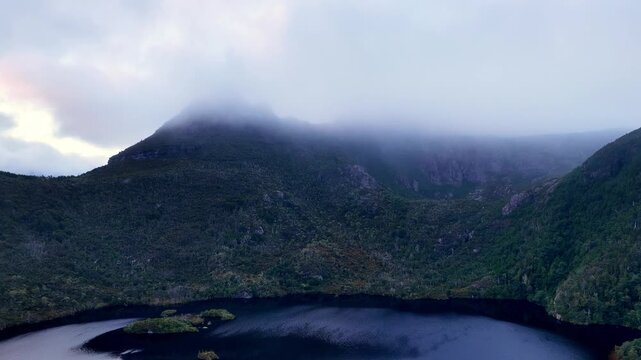 Back aerial flying high over Dove lake viewing Cradle Mountain in foggy morning weather.