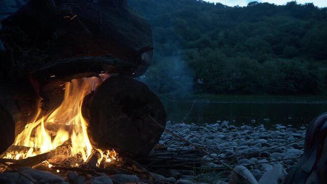 4K close-up video of a campfire placed on the left side of the frame, burning on stones beside the Drina River at sunset, with Bosnia and Herzegovina mountains visible from the Serbian side.
