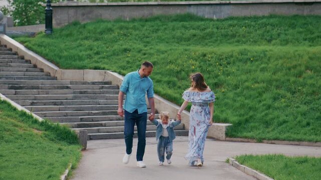 Family walks hand-in-hand through park path. Mother smiles, father laughs, child giggles along. Green grass and stone steps frame their joyful stroll. Ideal for