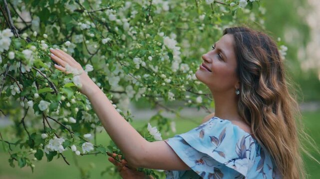 Woman smiles gently beneath blooming white blossoms. Her hands caress delicate flowers, feeling their softness. Sunlight filters through leaves, casting gentle patterns