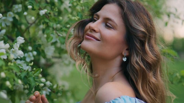 Soft light warms her face, eyes closed in peace. She brushes petals gently, smiling softly. Green branches frame her, white blossoms abundant. Spring's quiet joy radiates from her presence