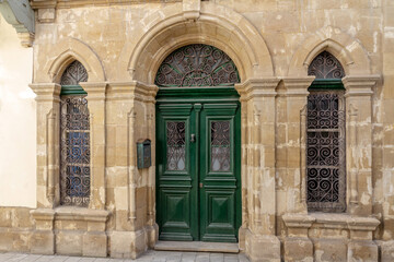 Fototapeta premium Vintage building facade made of limestone blocks with green wooden door. Traditional mediterranean architecture with arched portal and iron forged grates on windows.