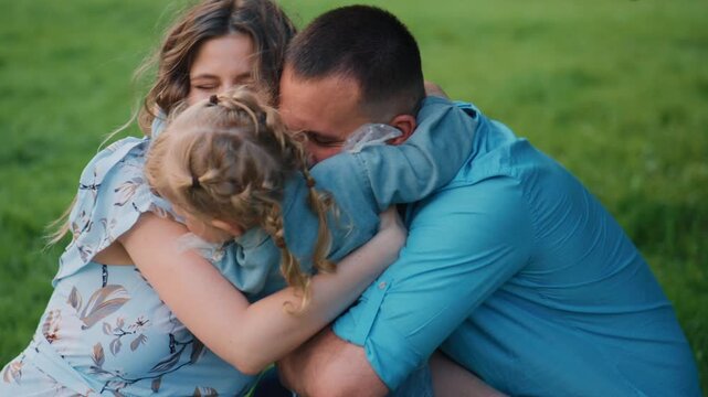 Family of three embraces on a sunlit green lawn. Mother and father kiss the child's head in affection. Child smiles warmly, surrounded by love